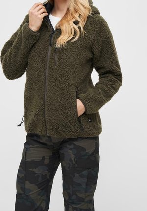 Woman wearing dark green fleece zip-up jacket with side and chest zip pockets and camouflage cargo pants, hand in jacket pocket.