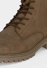 Botte marron clair à lacets avec motif texturé, coutures renforcées et semelle robuste à rainures profondes pour usage extérieur.