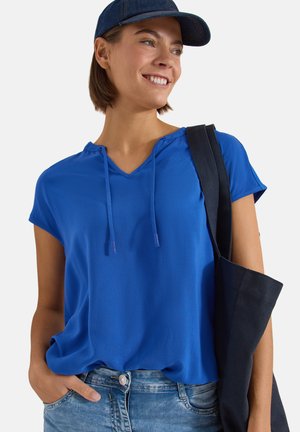 Jeune femme souriante, portant une casquette bleue et une chemise, avec un sac noir à l'épaule, une main dans la poche de son jean.