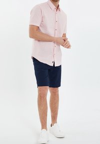 Kurzarm-Pinkhemd mit subtilen geometrischen Mustern, Holzknöpfen, navy Shorts und weißen Sneakern, die ein lässiges Ensemble bilden.