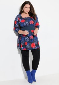 Túnica con estampado floral en azul marino, rojo y azul con mangas 3/4, cintura ajustada y combinada con leggings negros y botas de tacón azul.