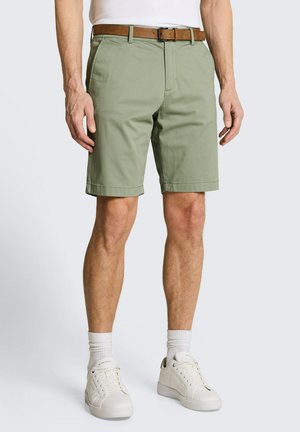 Mann trägt hellgrüne knielange Shorts mit braunem Gürtel, weißen Socken und weißen Sneakers und steht vor einem schlichten Hintergrund.