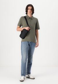 Olivenfarbener Polo mit kurzen Ärmeln und Reißverschlusskragen, hellblaue gerade Jeans und schwarze Umhängetasche. Das Modell trägt weiße Schuhe mit schwarzen Akzenten.