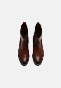 Une paire de bottes chelsea en cuir brun poli avec des bouts ronds et des tirettes, présentée sur un fond gris clair.