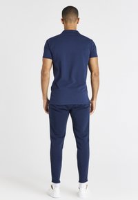 Mies, joka seisoo selkä kameraan päin, päällä tummansininen polo-paita, matching-housut, valkoiset sukat ja valkoiset lenkkarit valkoista taustaa vasten.