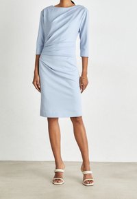 Vestido azul claro con mangas de tres cuartos, diseño drapeado en la cintura, dobladillo recto, hecho de una tela suave, combinado con sandalias beige.