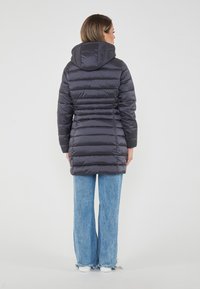 Donna che indossa un lungo giubbotto nero in piuma con cappuccio, abbinato a jeans blu a gamba larga e scarpe da ginnastica bianche, in piedi con le spalle rivolte verso di noi.