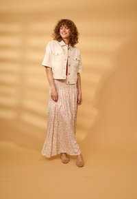 Femme aux cheveux bouclés portant une veste courte beige, une jupe longue fleurie et des chaussures tissées beiges, debout devant un fond marron clair.