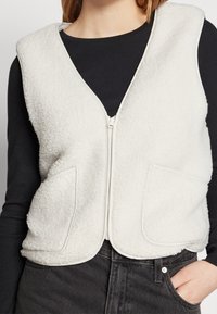 Vitt fleeceväst med dragkedja fram, som har två sidofickor och rundad halsringning. Materialet har en mjuk, strukturerad yta.