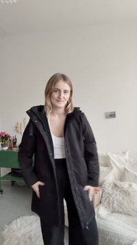 Črna odebeljena jakna s kapuco, opremljena z žepi na zadrgo in osrednjo zadrgo, nošena čez belo majico in črne hlače, v zaprtih prostorih.