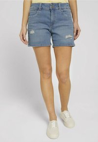 Donna che indossa pantaloni shorts in denim chiari strappati e sneakers bianche, in piedi contro uno sfondo chiaro e uniforme.