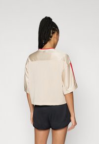 Top deportivo beige con espalda de malla, mangas cortas y cuello rojo. Combinado con pantalones cortos negros, mostrando un diseño cómodo y deportivo.