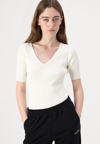 Weißes geripptes Strickoberteil mit kurzen Ärmeln und V-Ausschnitt, figurnaher Passform und dezenter Textur. Kombiniert mit schwarzen Jogginghosen mit Kordelzug.