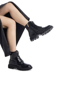 Botines de charol negro con parte superior texturizada, suela de goma con tacos, punta redonda y acentos de hardware en tono dorado. Cierre de cordones ajustado.