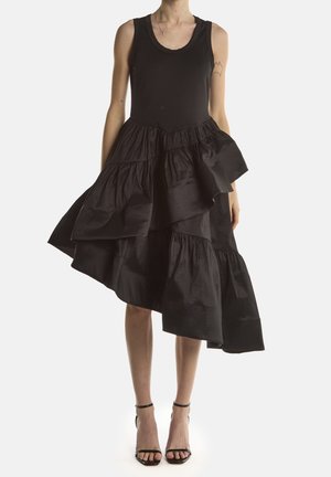 Kobieta ubrana w czarną, bezrękawową sukienkę z asymetryczną, warstwową spódnicą oraz czarne sandały na wysokim obcasie z paskiem wokół kostki, stojąca na białym tle.