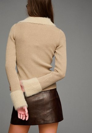 Beżowy sweter z prążkowanego dzianiny z futrzanymi mankietami i kołnierzem, połączony z brązową skórzaną spódnicą mini. Subtelna faktura i ciepłe odcienie w całej stylizacji.