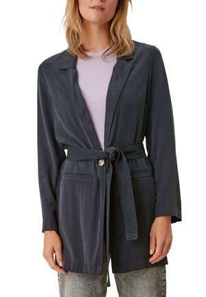 Frau trägt eine dunkel belted Jacke über einem lavendelfarbenen, gerippten Oberteil und hellgrauen Jeans, steht mit entspannt an den Seiten hängenden Händen.