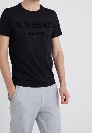 Homme portant un t-shirt noir à manches courtes avec le texte "ICEBERG depuis 1974", un pantalon de survêtement gris clair, debout devant un fond clair uni.