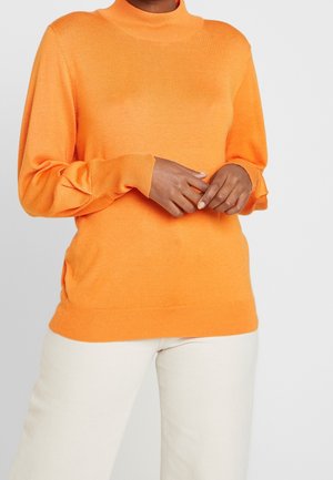 Person wearing a bright orange long-sleeve sweater with gathered sleeves and white pants, standing against a plain light background.