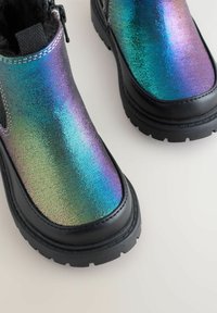 Stivali neri dal design massiccio con tomaia iridescente e brillante che mostra sfumature di blu, verde e viola, con dettagli di cucitura ben visibili.