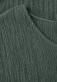 Tessuto verde texturizzato con righe verticali e una sezione colletto, caratterizzato da una cucitura liscia lungo il bordo. Materiale morbido e leggero.