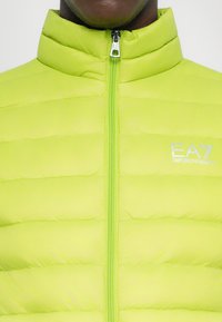Zelená puffer bunda s vysokým golierom, s vertikálnym prešívaním, plným zipsom a reflexným logom EA7 na ľavom hrudníku.