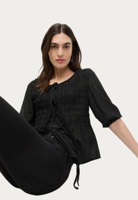 Schwarze Bluse mit Rüschendetails, rundem Ausschnitt und Bindebändern. Lange, leicht gepuffte Ärmel; leichtes, strukturiertes Gewebe.