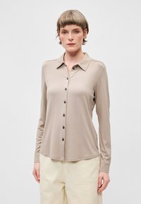 Svilena bež košulja dugih rukava s kragnom, izrađena od mekog materijala, sa tamnim gumbima i opuštenim krojem.
