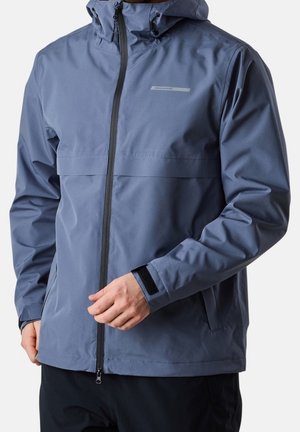 Man draagt een blauwe waterdichte jas met capuchon, voorrits en verstelbare polsbanden, staand tegen een effen achtergrond.