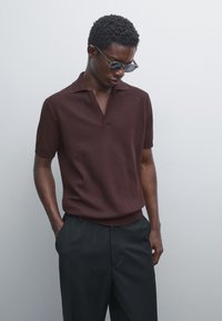 Mand iført mørke solbriller, kortærmet brun strikket polo-shirt og sorte bukser, står med hænderne i lommerne mod en ensfarvet baggrund.