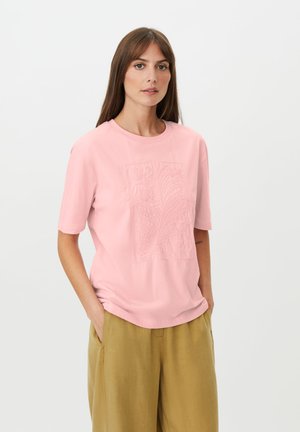 Kvinde med langt brunt hår iført en lyserød tekstureret t-shirt og løse sennepsgule bukser, stående med hænderne i lommerne mod en hvid baggrund.