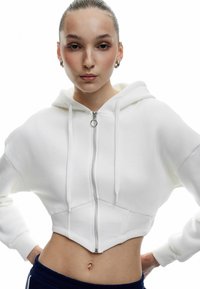 Weißer, verkürzter Hoodie mit einem Reißverschluss und einer verstellbaren Kapuze. Aus strukturiertem Stoff gefertigt, bietet er eine figurbetonte Passform, die die Taille betont.