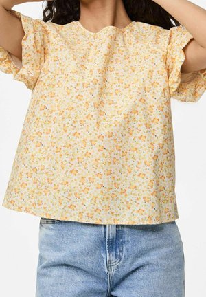 Woman wearing a loose, short-sleeved yellow floral blouse paired with light blue high-waisted jeans, arms raised above her head.