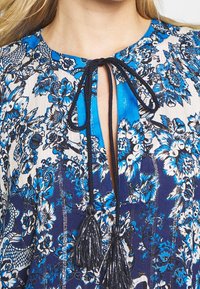 Närbild av en blå och vit blommig blus med svart tofsband vid halsringningen och vertikalt sprund som visar hud.