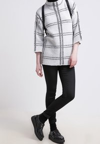 Přesměrně šedý a bílý kostkovaný svetr s černými akcenty, doplněný o černé přiléhavé kalhoty a texturované černé boty s kovovým prvkem.