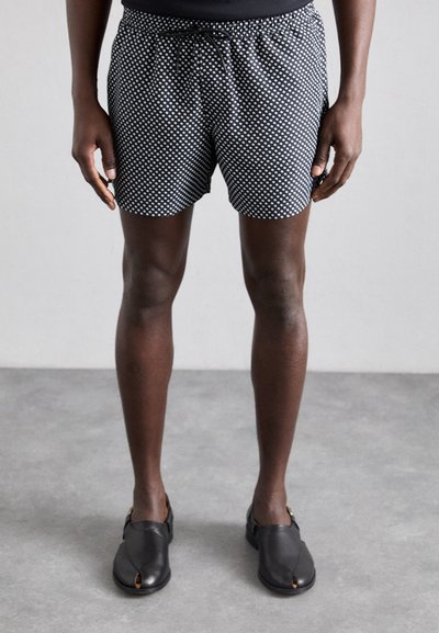 Shorts noirs à pois blancs, avec une taille élastique et un cordon de serrage. Portés avec des chaussures noires à enfiler.