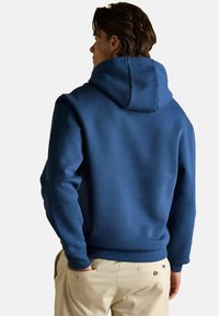 Tumesinine kapuutsiga sviiter, valmistatud pehmest kangast, millel on reguleeritav kapuuts ja ribakujulised varrukad. Mudel kannab beeže pükse, millel on tagatasku.