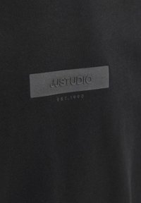 Črna bombažna majica s teksturiranim sivim označevalcem, na katerem je v preprostem fontu vtisnjeno "JJSTUDIO" in "EST. 1990". Gladek material.