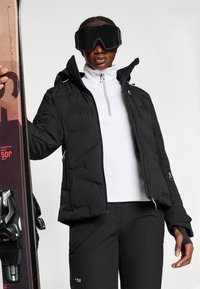 Melna pūkainā jaka ar augstu apkakli, ar rāvējslēdzēju kabatām un rozā akcentiem, apvienota ar baltu garo piedurkņu topu un melniem sniega biksēm.