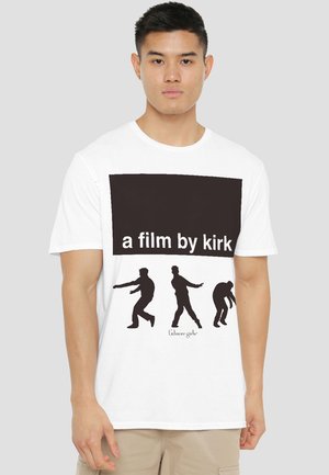 Young man wearing a white t-shirt with black text "a film by kirk" and silhouettes of three walking figures below, with "Gilmore girls" text.