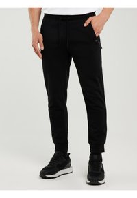 Mees, kes kannab musta tõmblukuga vöökohaga dressipükse ja musti spordijalatseid, seisab ühevärvilise heleda tausta ees.