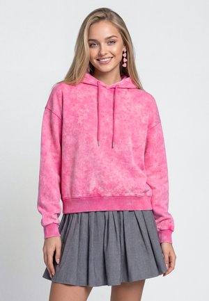 Jonge vrouw die lacht, draagt een roze tie-dye hoodie met trekkoorden en een grijze geplooide rok, staand tegen een effen achtergrond.