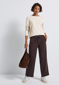 Femme se tenant debout, portant un haut à manches longues crème, un pantalon à cordon de serrage marron foncé, des baskets blanches et un sac à main en daim marron foncé.