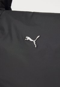 Zwarte stof met een gladde textuur heeft een zilveren Puma-logo op de zijkant, met een ontwerp van een springende kat in een metallic afwerking.