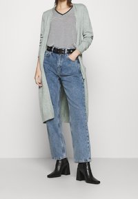 Žena v šedém dlouhém kardiganu, černo-bílém pruhovaném tričku, vysokých modrých džínách s černým páskem a černých kožených kotníkových botách na podpatku.