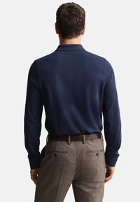 Tmavomodré polokošeľa s dlhými rukávmi a jemnou textúrou, v kombinácii s hnedými tweedovými nohavicami s klasickým vzorom psieho zub a zadnými vreckami.