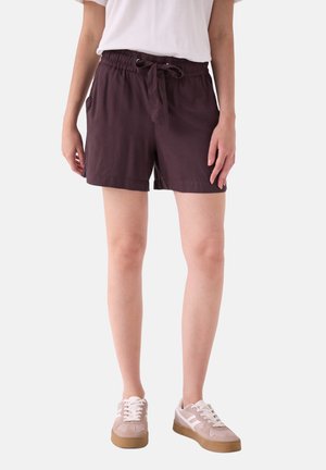 Person iført mørkelilla shorts med snøret talje, hvid t-shirt og beige og hvide sneakers, stående mod en ensfarvet baggrund.