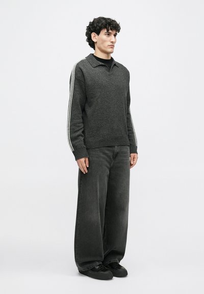 Grijze gebreide trui met een kraag en drie witte strepen op elke schouder, in combinatie met brede zwarte jeans en donkere schoenen.