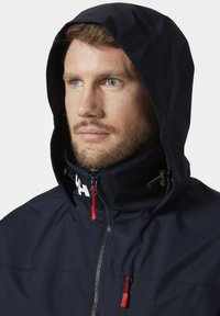 Marineblauwe hoodie met een hoge kraag, ritsluiting, rode ritsen en een logo bij de hals. Gladde textuur met een modern ontwerp.