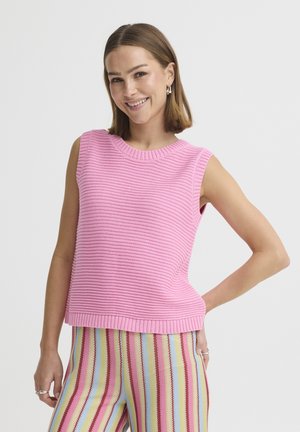 Uśmiechnięta kobieta o krótkich brązowych włosach, ubrana w różowy bezrękawnik dzianinowy i wielokolorowe spodnie w pionowe paski, pozująca z jedną ręką na biodrze.